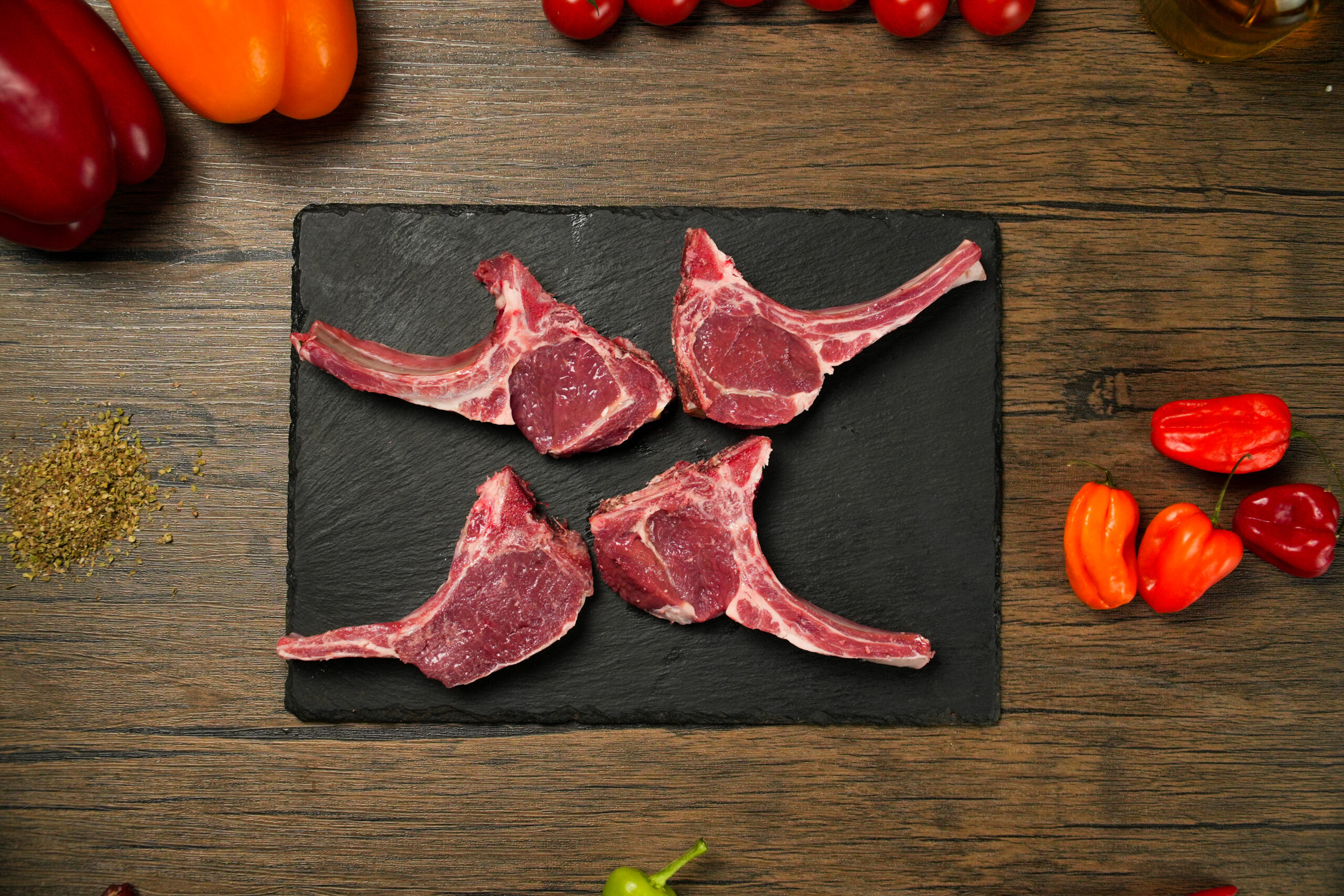 Raw halal lamb chops on a black slate board, four bone-in lamb cutlets with long rib bones and visible marbling, arranged neatly on a rustic wooden surface, surrounded by colorful ingredients including orange and red bell peppers, cherry tomatoes, and small chili peppers, with scattered dried herbs, gourmet butcher presentation for grilling and cooking