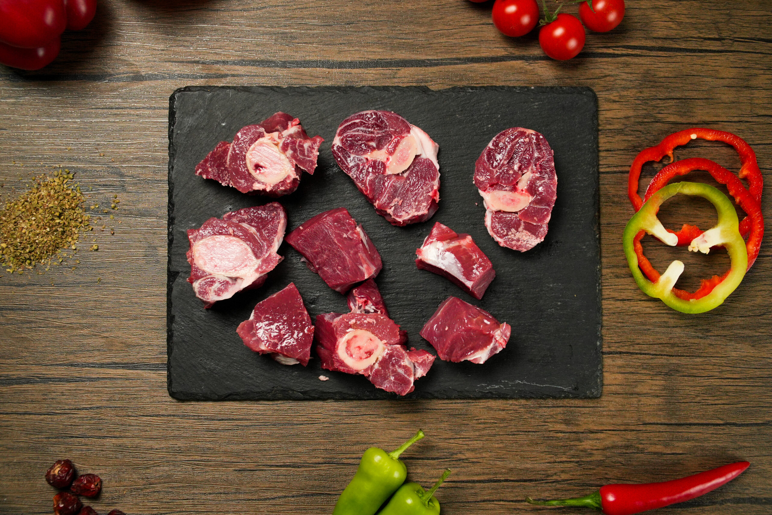 Raw mutton bone-in pieces (mixed cuts including marrow bones and diced chunks) arranged on a black slate board, showing deep red meat with visible bone cross-sections and creamy marrow centers. Surrounded by fresh ingredients such as sliced red and green bell peppers, cherry tomatoes, chili peppers, dried berries, and scattered herbs on a rustic wooden surface, ideal for halal cooking, slow-cooked stews, curries, and traditional meat preparation.