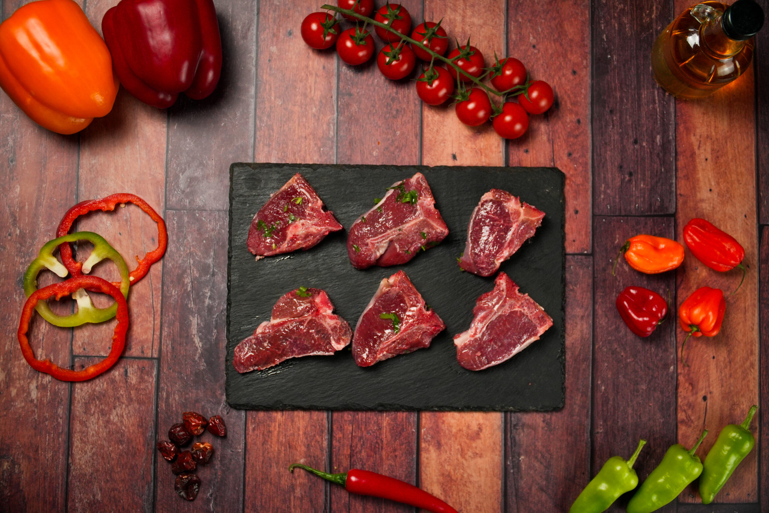Raw halal lamb chops arranged on a black slate serving board, six fresh bone-in meat cuts with light marbling and parsley garnish, surrounded by colorful ingredients including sliced bell peppers, whole chili peppers, cherry tomatoes on the vine, and a glass bottle of oil, rustic wooden background, gourmet butcher presentation for cooking and grilling