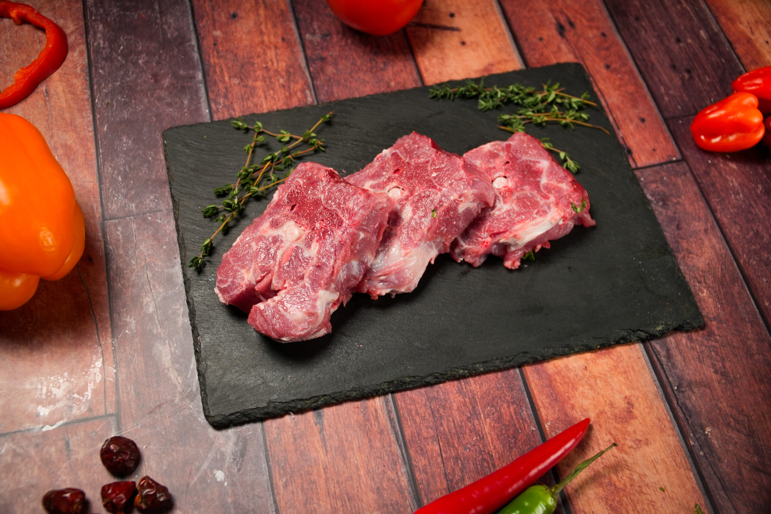 Raw lamb neck chops on a black slate board, three bone-in lamb cuts with visible marrow and connective tissue, fresh halal meat with light marbling, garnished with thyme sprigs, surrounded by colorful ingredients including chili peppers and bell peppers on a rustic wooden background, ideal for slow cooking, braising, and rich stew recipes