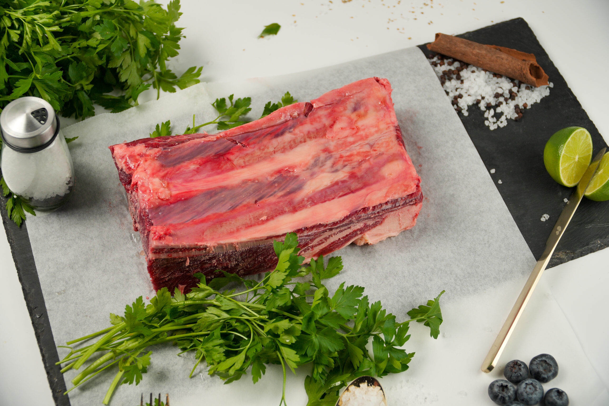 A raw rack of beef ribs on parchment paper, surrounded by fresh parsley, coarse salt, a cinnamon stick, lime halves, black pepper, and blueberries on a black slate board.