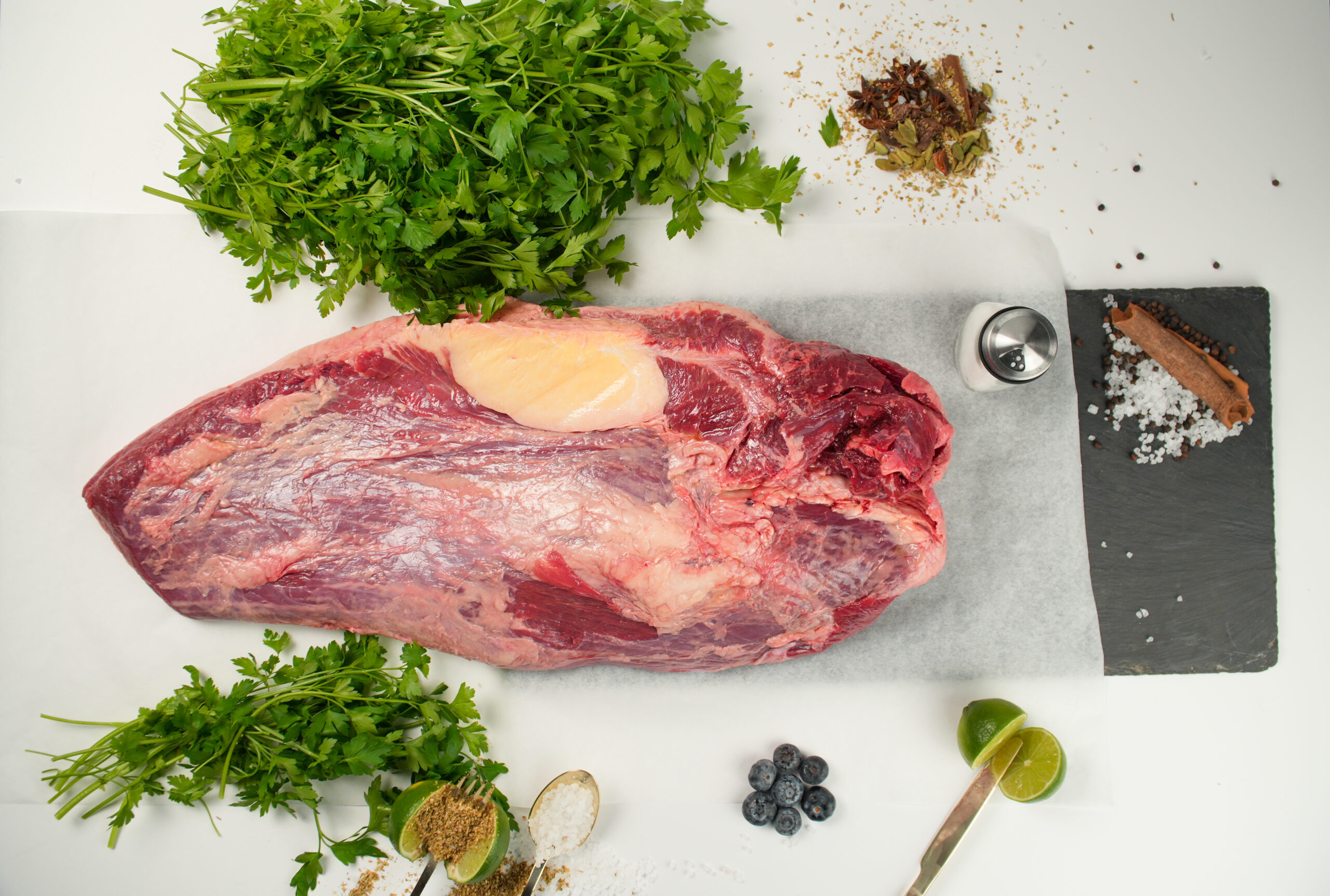 A large, raw cut of beef sits on parchment paper, surrounded by fresh herbs, spices, coarse salt, blueberries, and lime halves on a white surface.
