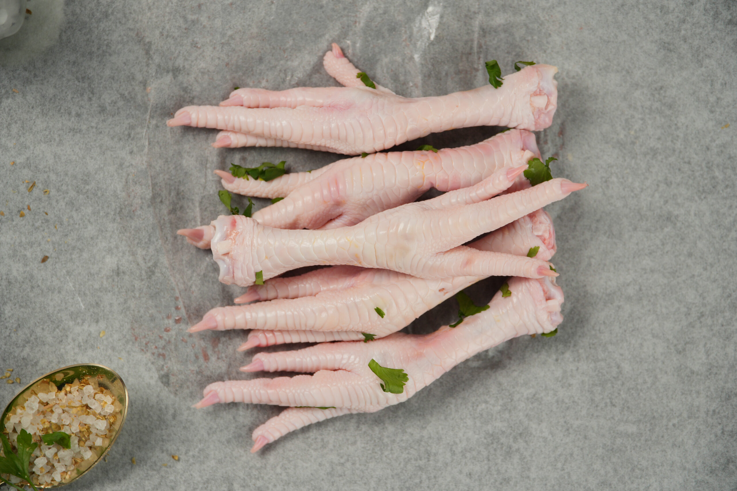 Raw halal chicken feet on parchment paper, fresh poultry offal with visible texture, garnished with herbs and seasoning in a clean preparation setting