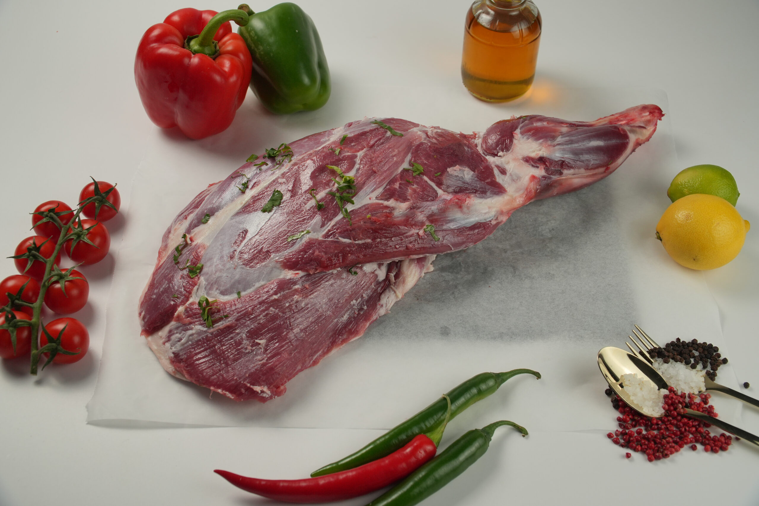 Raw whole mutton leg (bone-in) displayed on parchment paper, featuring deep red lean meat with light fat trimming and visible muscle structure, garnished with chopped fresh herbs. Surrounded by fresh ingredients including cherry tomatoes on the vine, red and green bell peppers, lemon, lime, chili peppers, and a glass bottle of oil, with coarse salt and peppercorns in gold utensils on a clean white background—ideal for halal butchery, roasting cuts, and premium meat presentation.