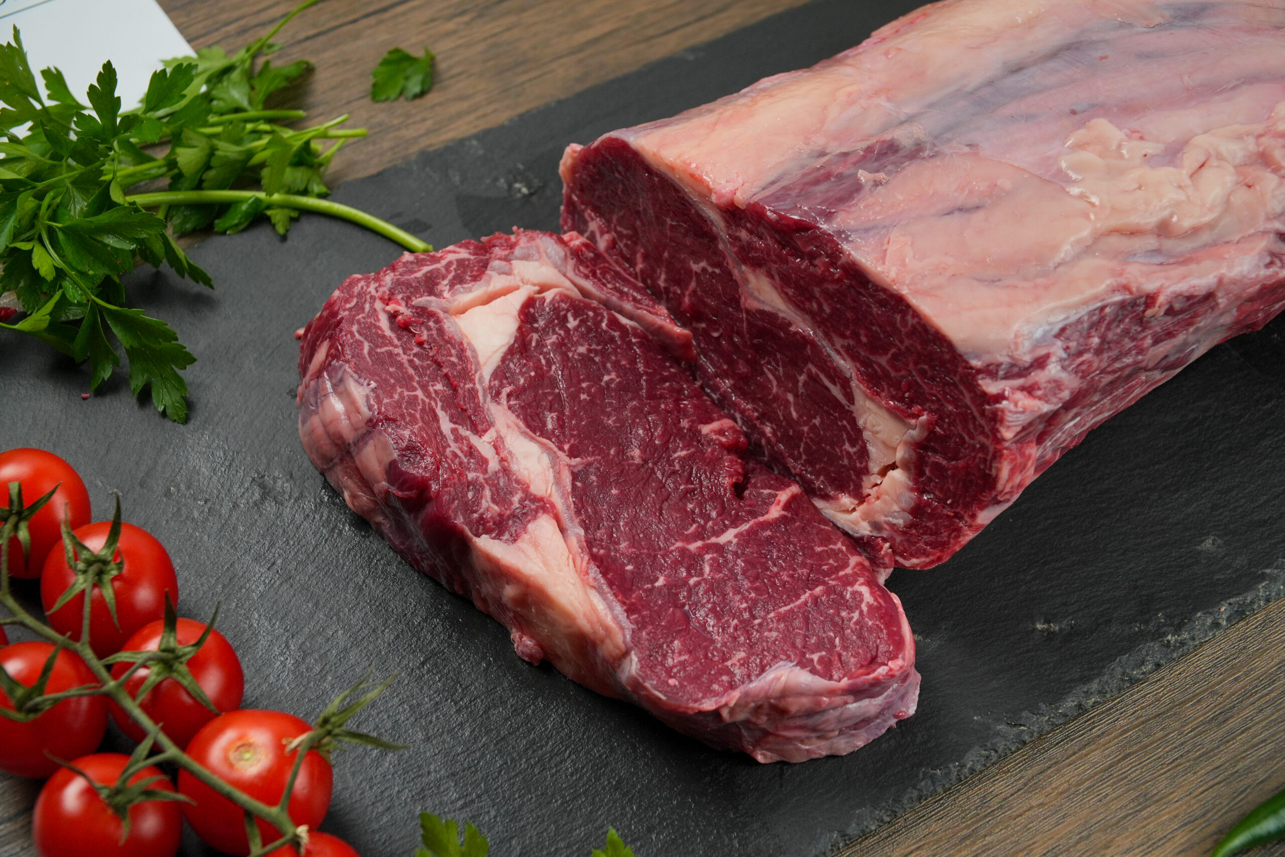 Raw marbled beef ribeye roast with thick-cut steaks on black slate board, fresh cherry tomatoes and parsley garnish, close-up of premium halal beef with visible fat cap and rich red texture, butcher-style presentation for gourmet cooking and steak preparation