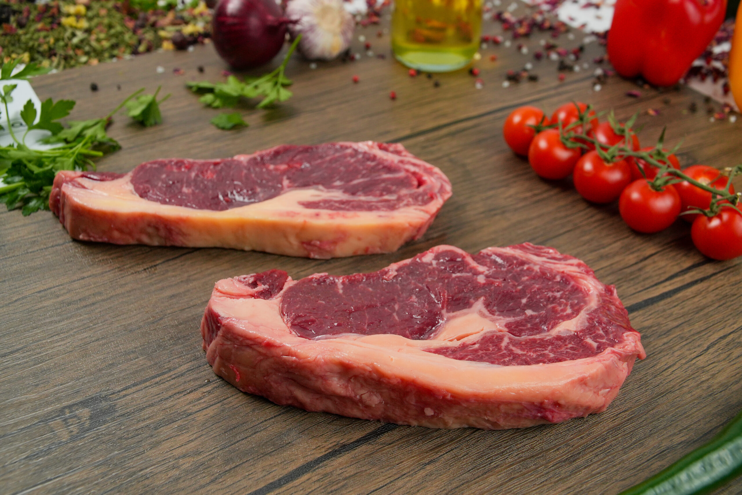 Raw beef ribeye steaks with thick fat cap on wooden cutting board, fresh cherry tomatoes on vine, garlic, herbs, and spices in background, premium halal beef cuts with rich marbling, butcher-style presentation for grilling and steak cooking, rustic kitchen food photography