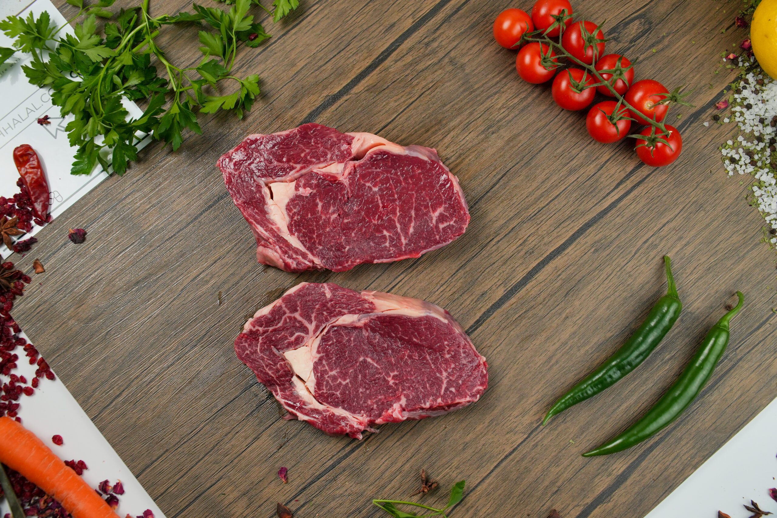 Two raw beef steaks on a wooden surface, surrounded by cherry tomatoes on the vine, parsley, green chilies, sea salt, carrot, a lemon, and assorted spices.