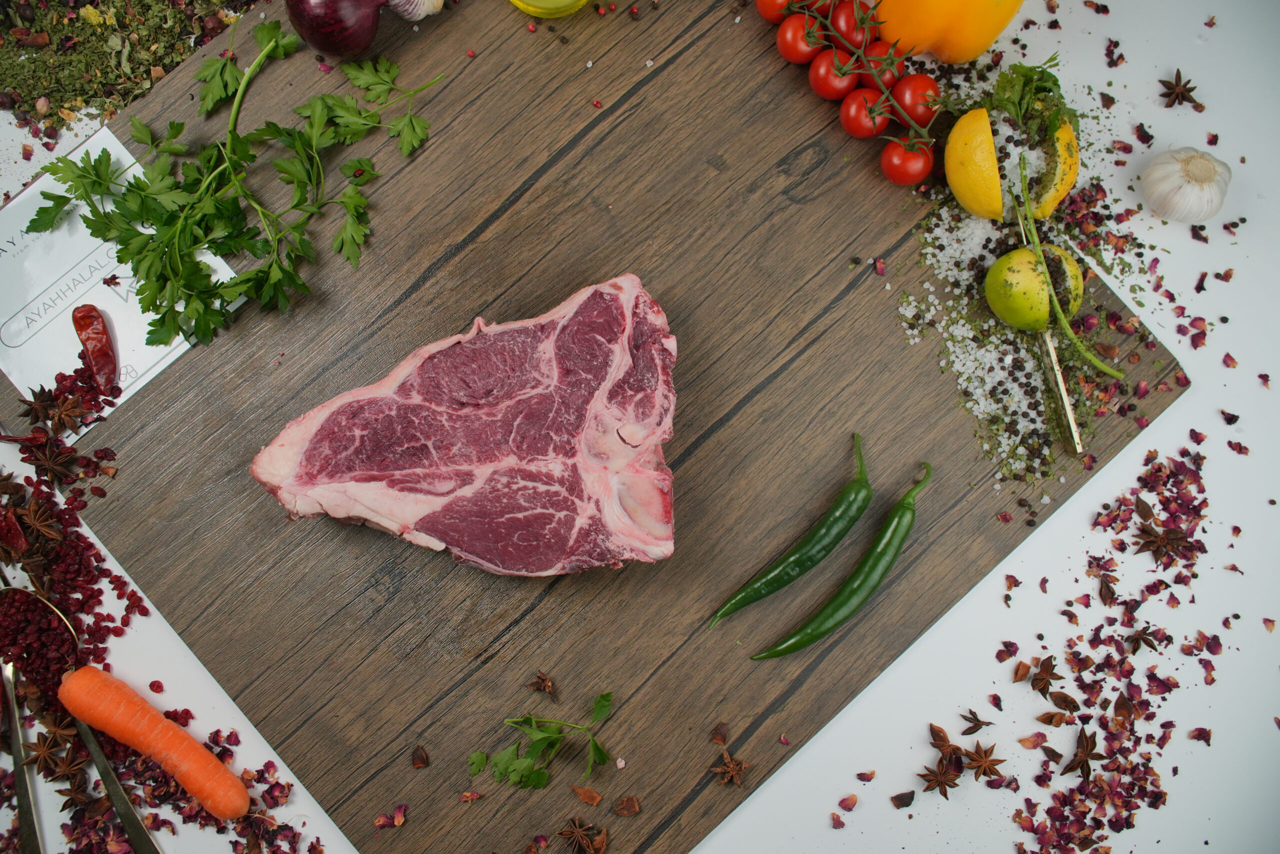 Raw beef T-bone steak on wooden cutting board, marbled red meat with visible fat and bone, surrounded by fresh ingredients including green chili peppers, cherry tomatoes, parsley, lemon wedges, garlic, carrot, and scattered spices, rustic food styling with Ayah Halal branding partially visible, butcher-quality halal beef concept