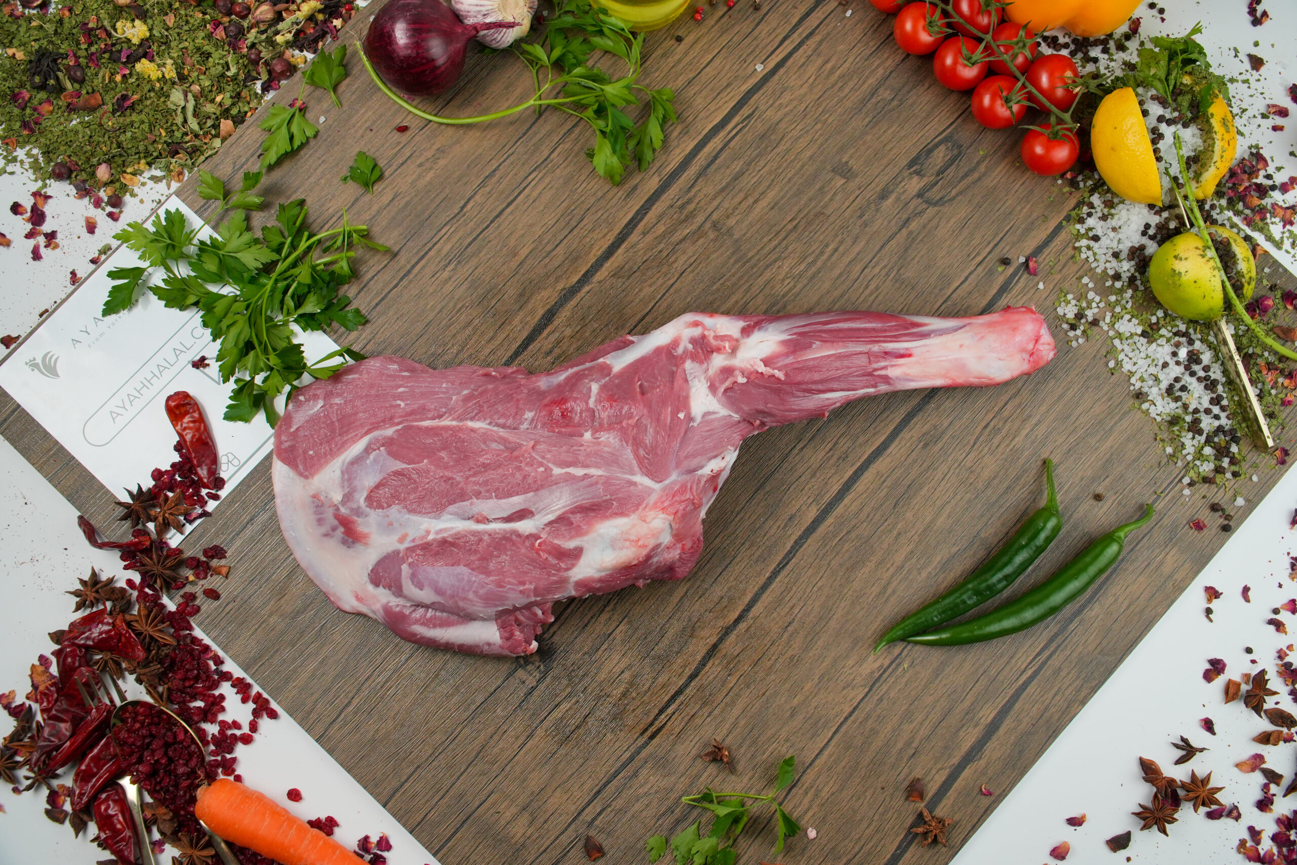 Raw whole lamb leg (bone-in) displayed on a wooden board, fresh red meat with visible fat marbling, surrounded by herbs, spices, chili peppers, garlic, cherry tomatoes, lemon, and seasoning ingredients for gourmet cooking preparation