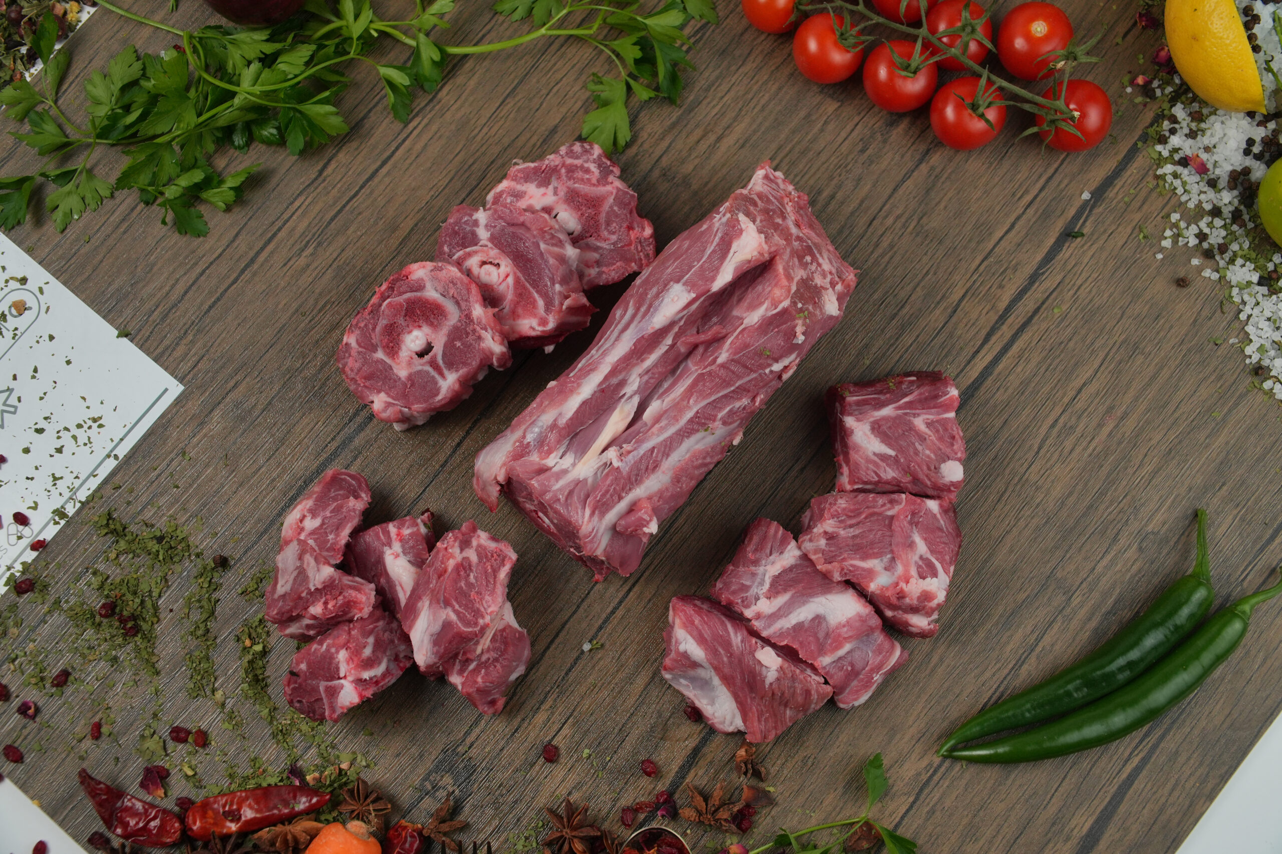 Raw halal lamb cuts including neck, ribs, and diced pieces on a wooden surface, fresh bone-in meat with herbs, spices, and vegetables for cooking