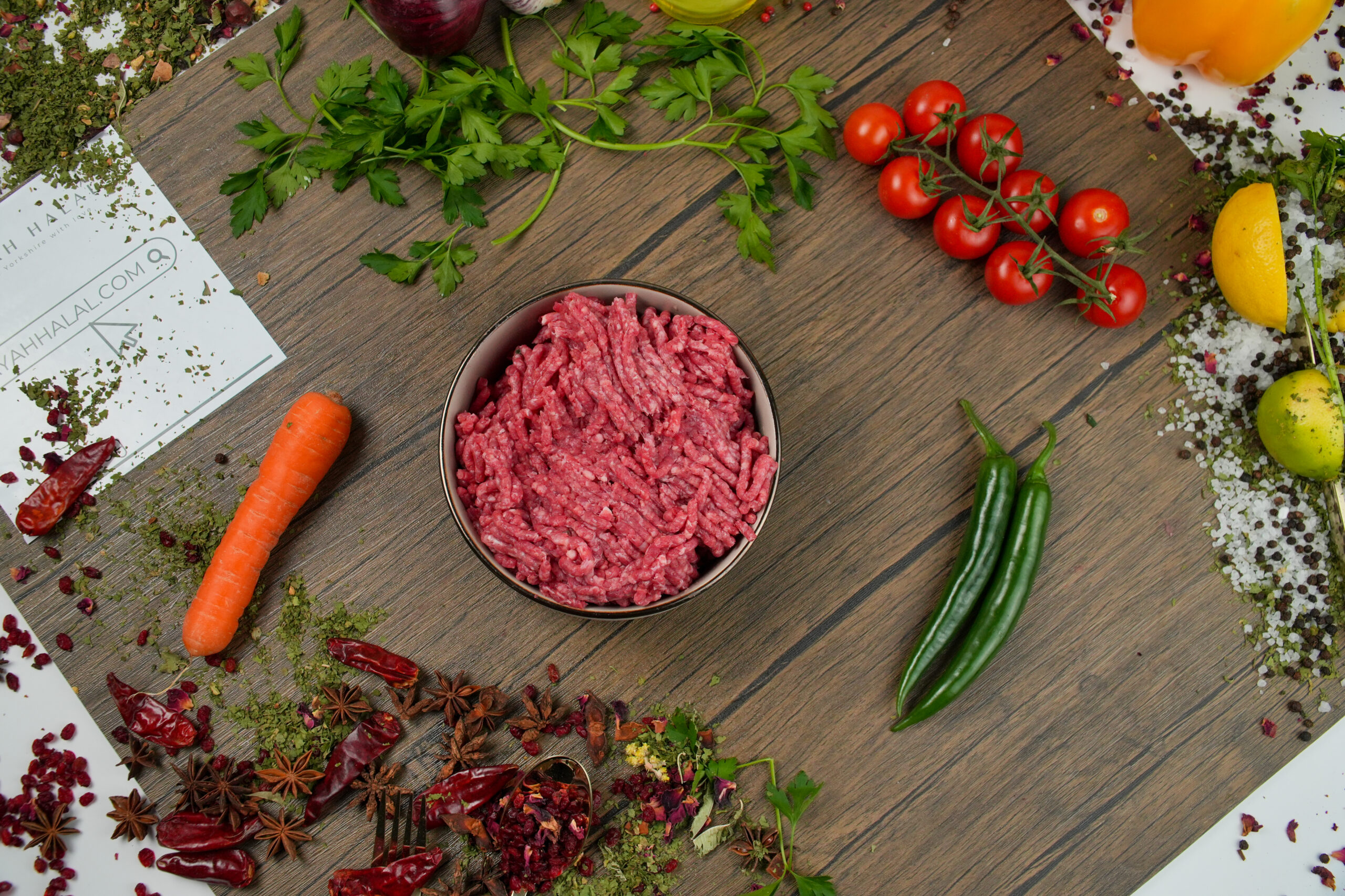Raw ground beef mince in a bowl on a wooden surface, fresh minced red meat with visible texture, surrounded by parsley, cherry tomatoes, green chili peppers, carrot, garlic, dried spices, and herbs, with partial “Ayah Halal” branding visible for food preparation and cooking concept