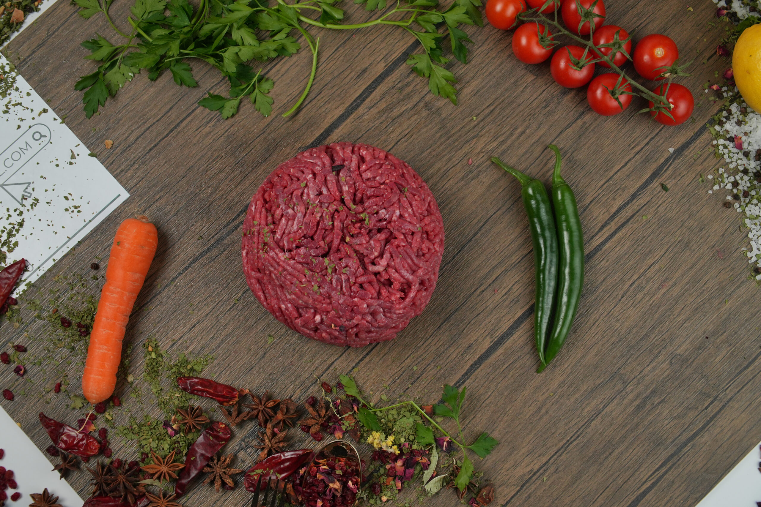 Raw ground beef mince patty centered on a wooden surface, fresh minced meat with visible texture, surrounded by green chili peppers, cherry tomatoes, carrot, parsley, dried spices, and herbs for cooking and meal preparation concept