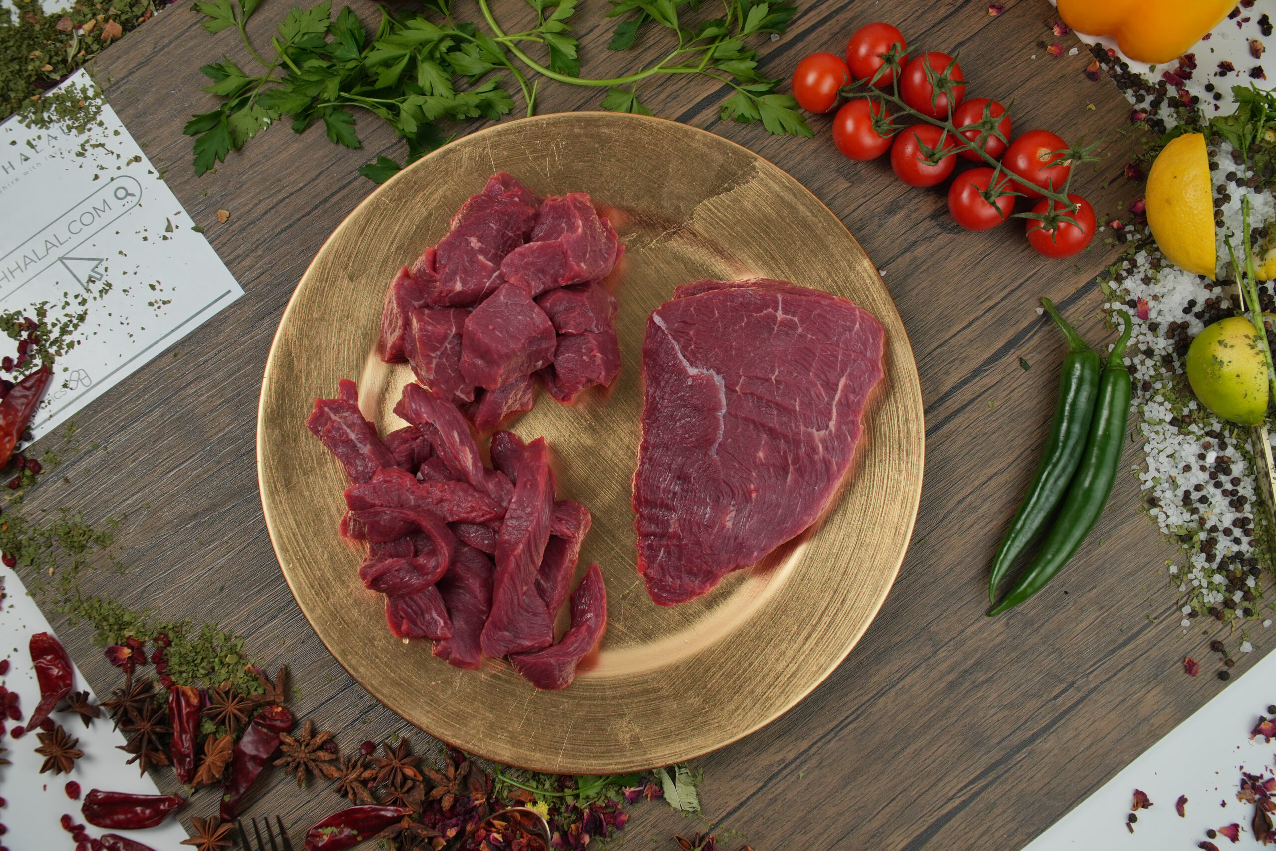 Raw halal beef assortment on gold plate, featuring diced beef cubes, sliced beef strips, and whole lean beef cut, surrounded by fresh ingredients including cherry tomatoes on the vine, green chili peppers, parsley, lemon, garlic, and mixed spices, rustic wooden background with visible “AYAHHALAL.COM” branding, premium butcher-style beef preparation for cooking recipes
