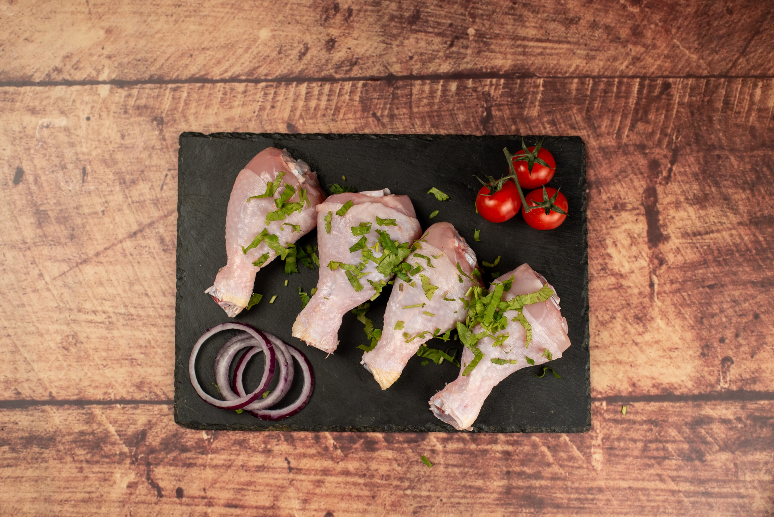 Raw halal chicken drumsticks on a slate board, fresh poultry with chopped herbs, onion slices, and cherry tomatoes on a rustic wooden background