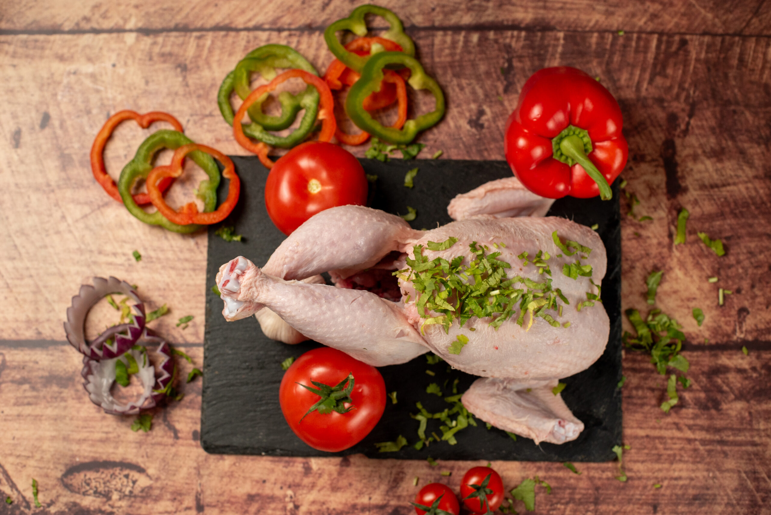 Whole raw halal chicken on a slate board, fresh poultry with herbs, tomatoes, peppers, and onion on a rustic wooden background