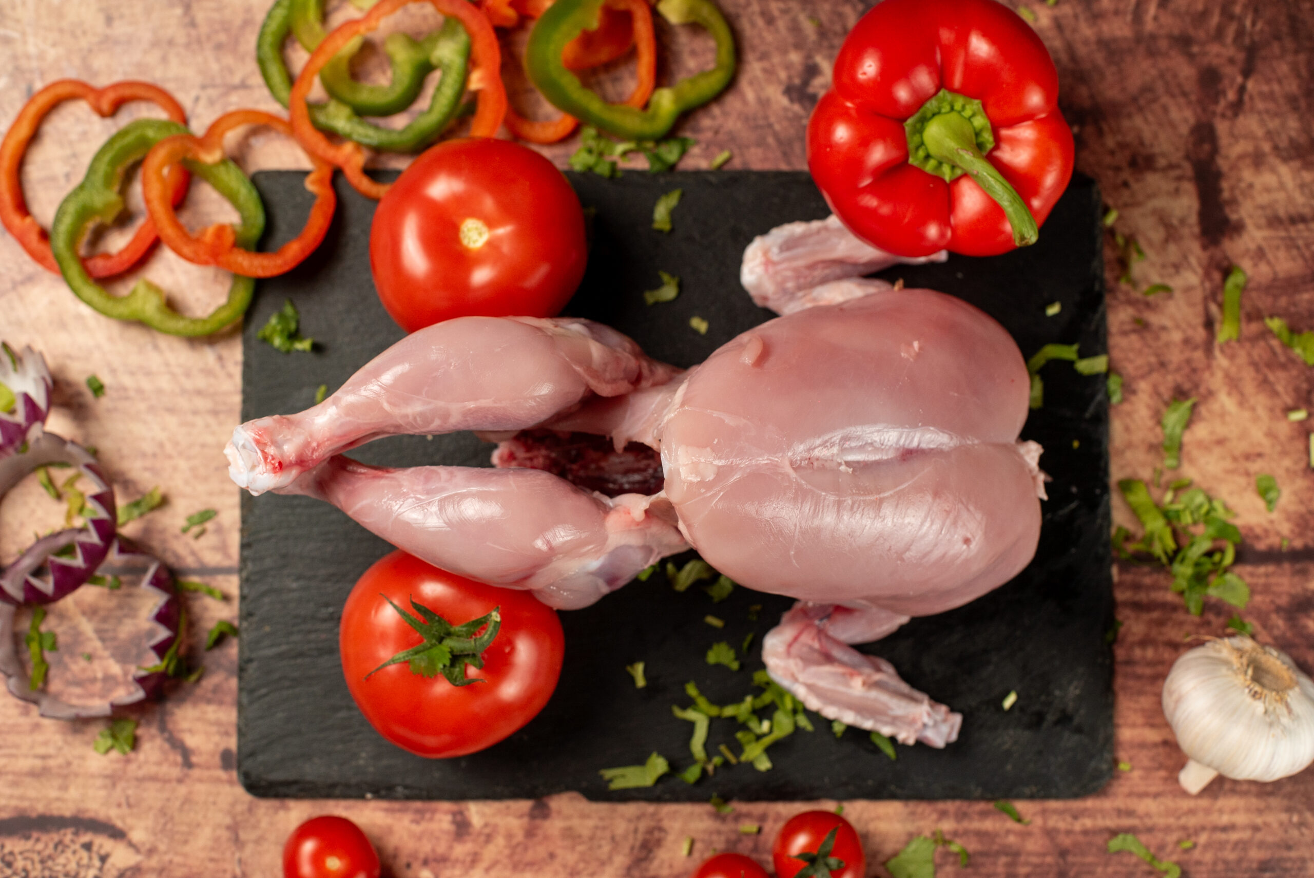 Whole raw halal chicken on a slate board, fresh poultry with tomatoes, peppers, garlic, and herbs on a rustic wooden surface