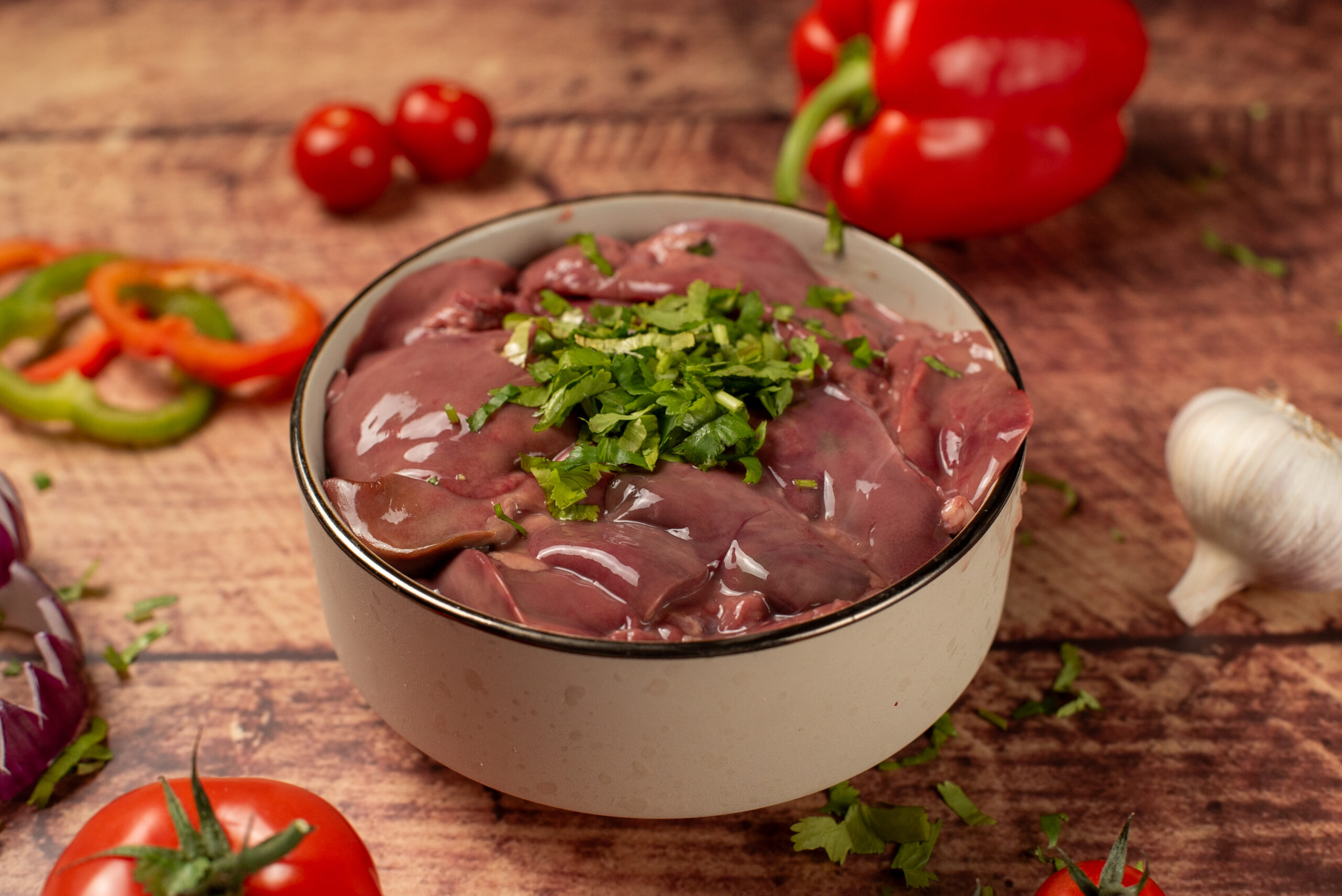 Raw halal chicken liver in a bowl, fresh poultry offal with herbs, surrounded by vegetables and spices on a rustic wooden surface