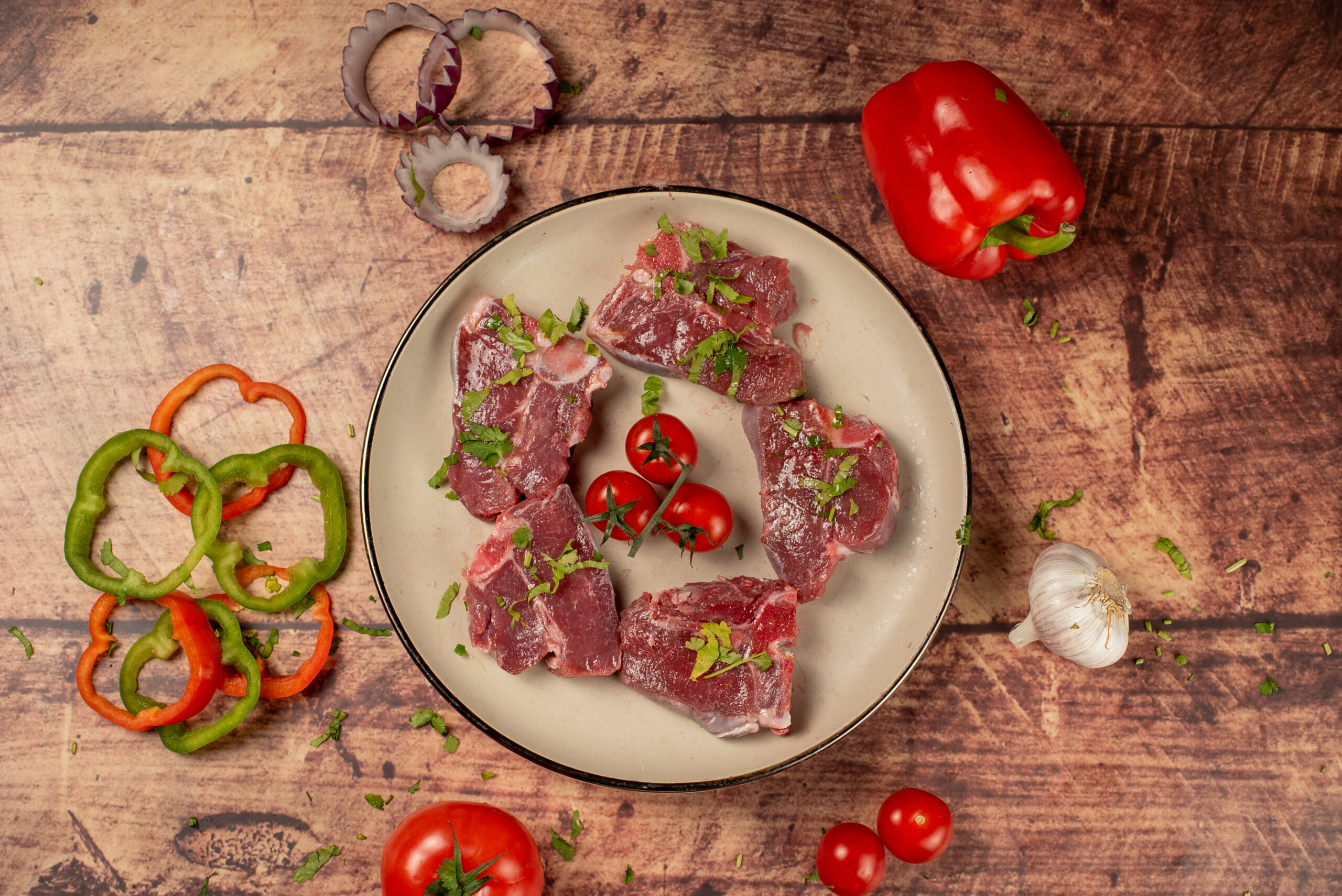 Raw halal lamb chops arranged in a bowl, fresh bone-in meat with herbs, tomatoes, peppers, and garlic on a rustic wooden background