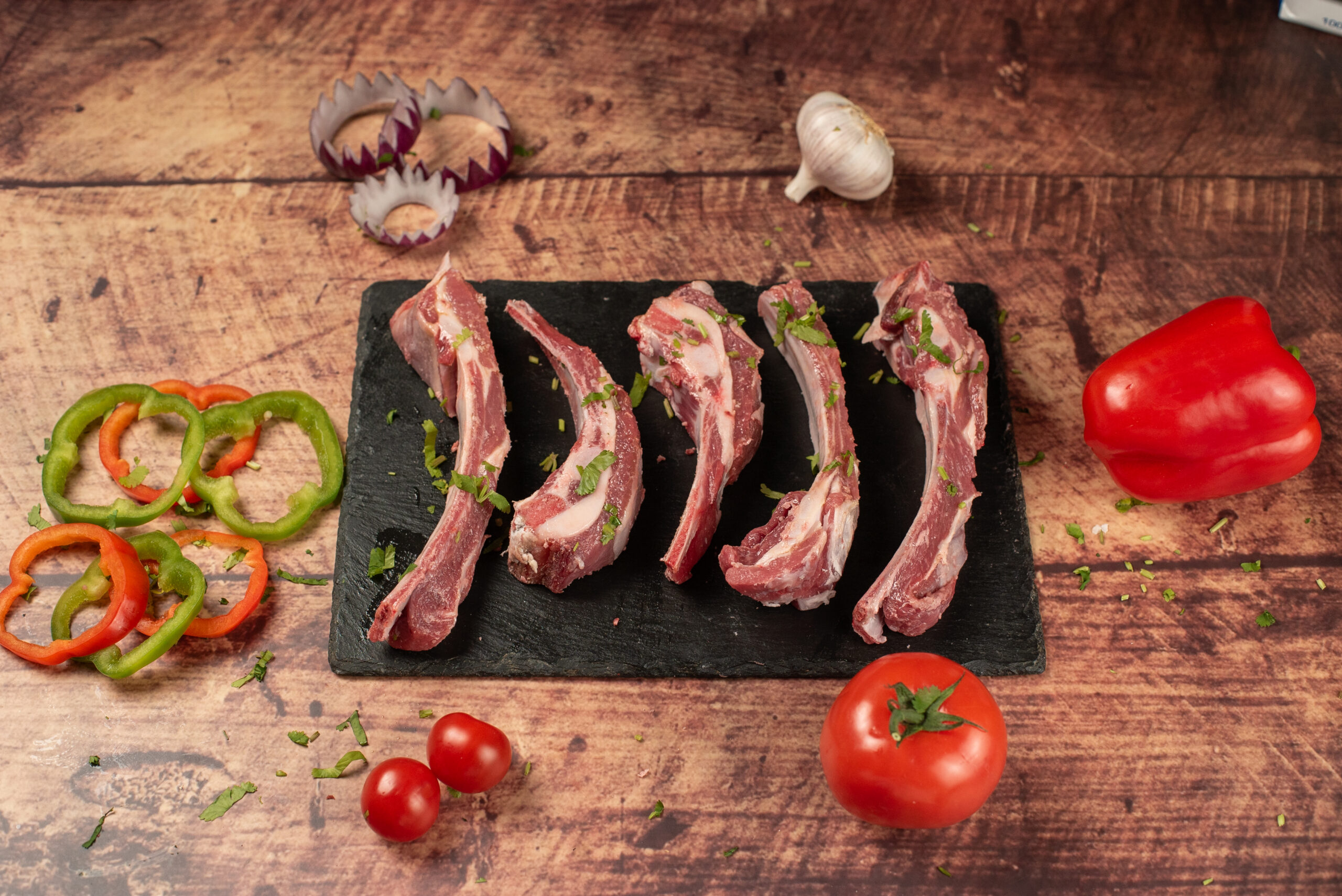 Raw halal lamb ribs on a slate board, fresh bone-in rib cuts with herbs, peppers, and vegetables on a rustic wooden background