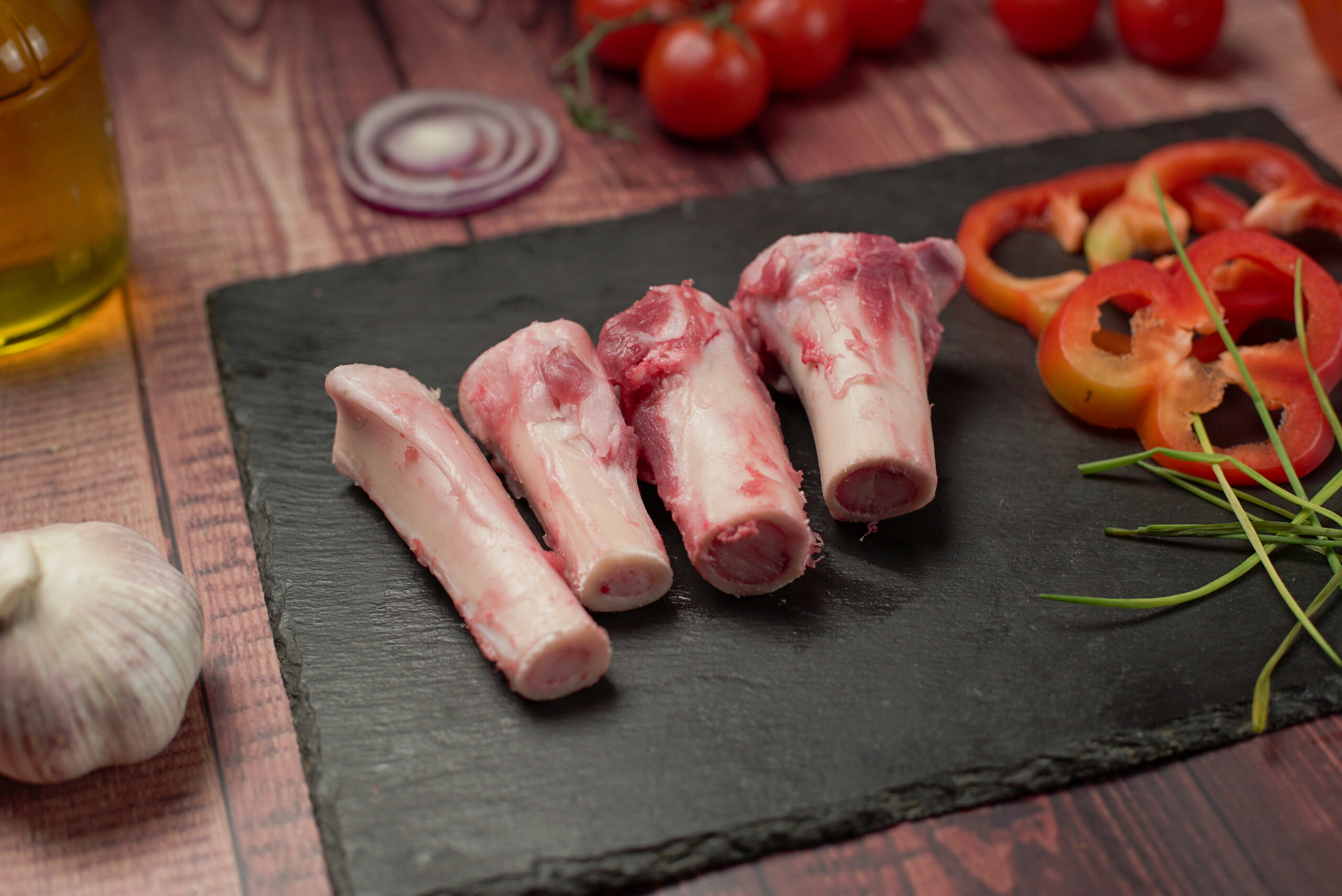 Raw halal lamb bones cut into sections on a slate board, fresh marrow bones with garlic, peppers, and herbs on a rustic wooden background
