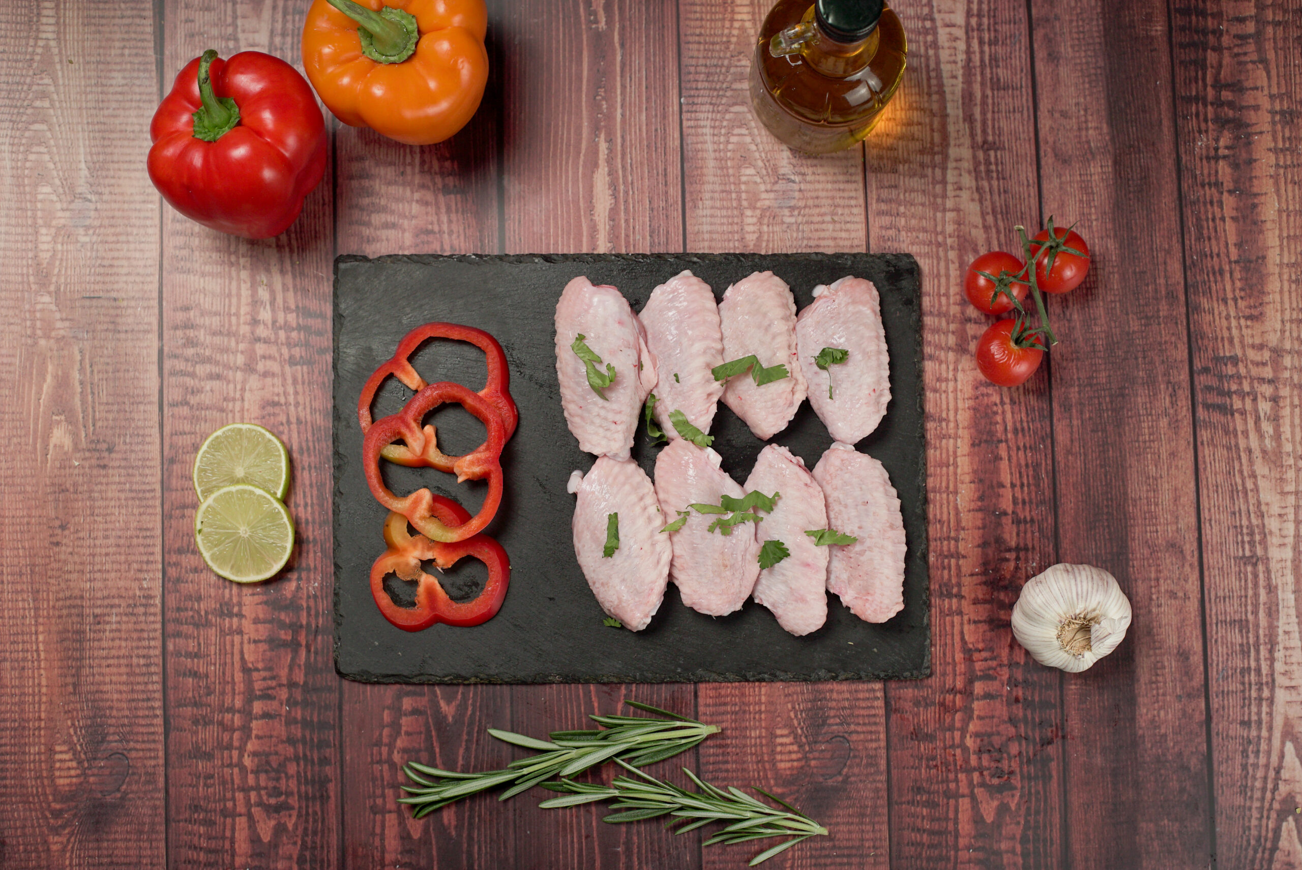 Raw halal chicken wing tips on a slate board, fresh poultry cuts with sliced peppers, herbs, lime, and garlic on a rustic wooden background