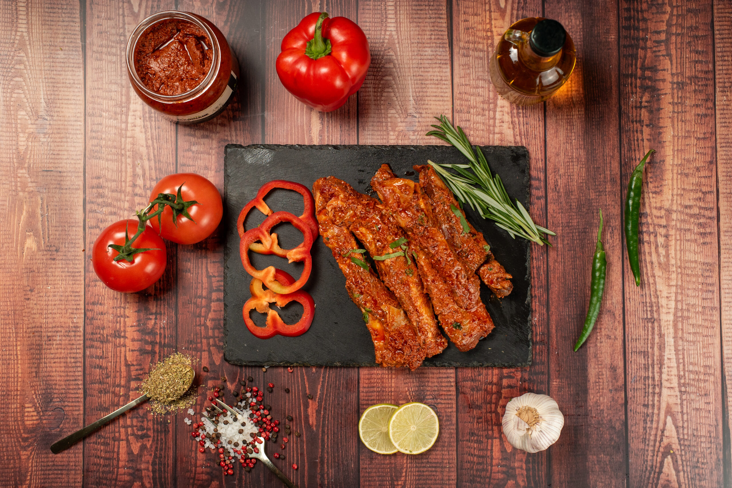 Grilled ribs on slate with rosemary, red peppers, tomatoes, spices, and lemon slices on a wooden table.