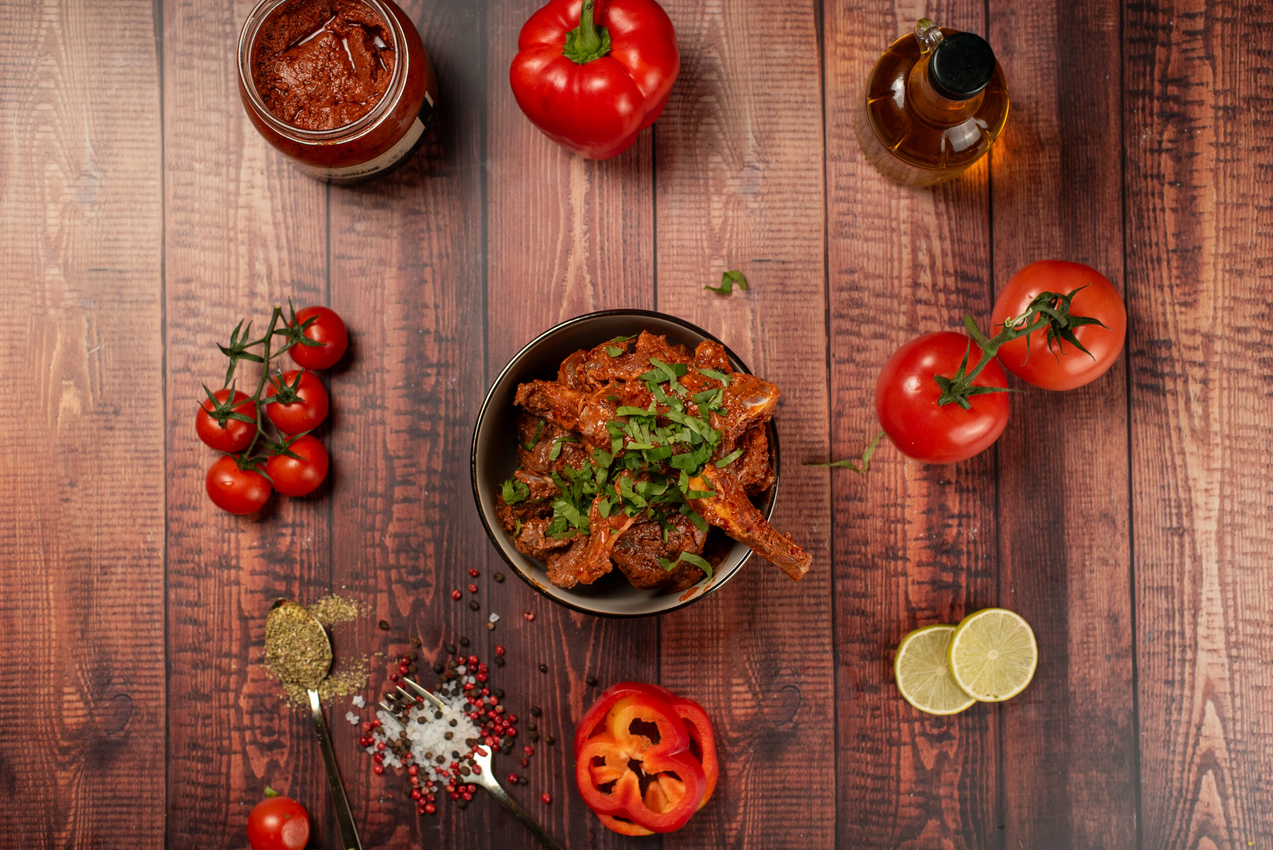 A bowl of seasoned meat garnished with herbs sits on a wooden table surrounded by tomatoes, red pepper, lime slices, spices, a jar of sauce, and a bottle of oil.