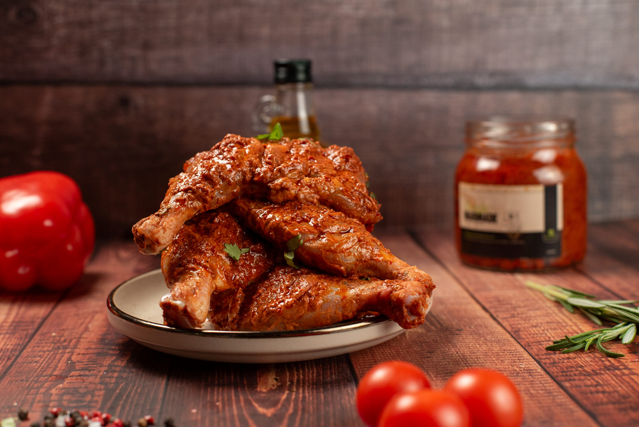 Spicy marinated chicken drumsticks on a plate with red pepper, cherry tomatoes, and a jar of sauce on a wooden table.