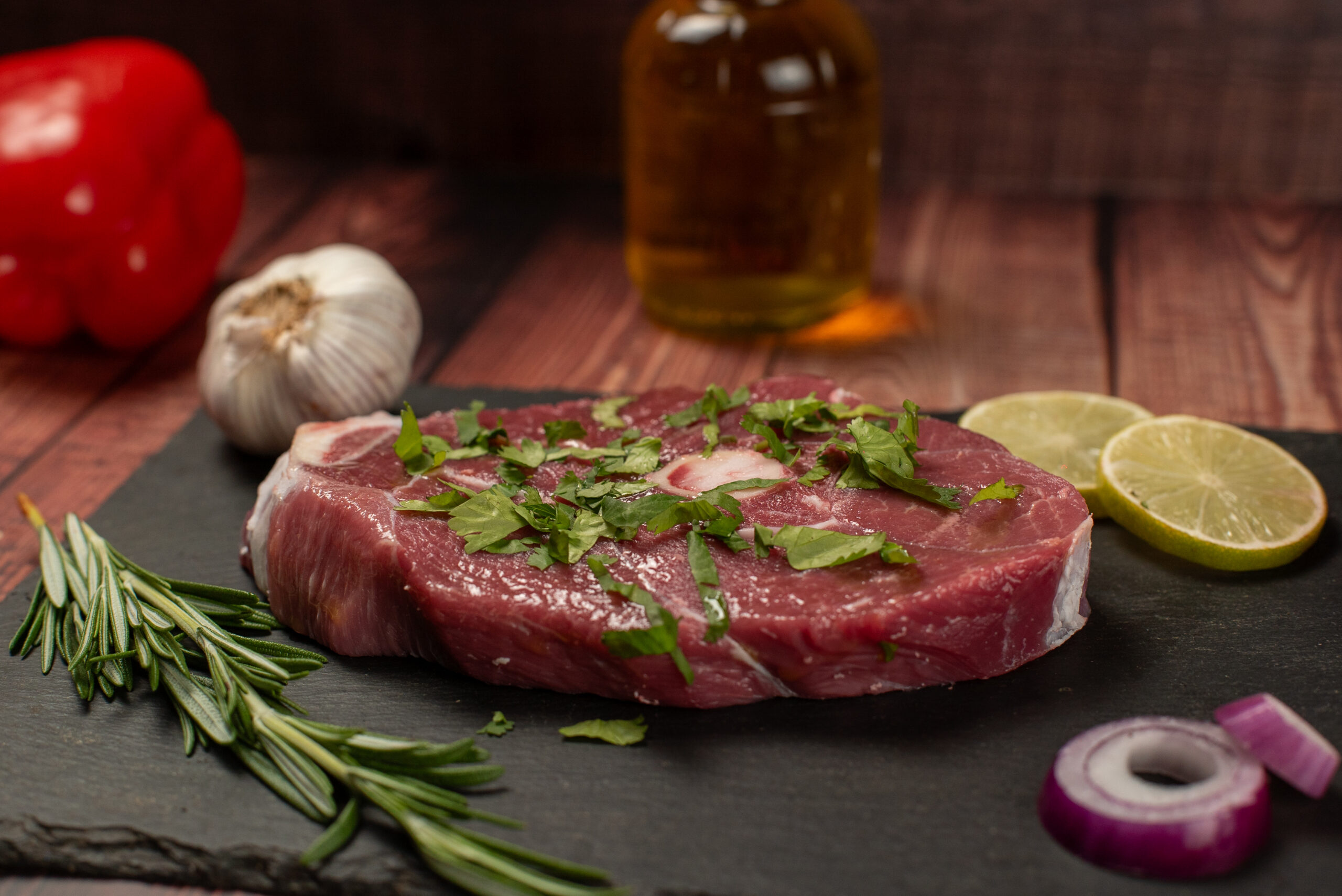 Raw mutton steak (bone-in round cut) placed on a black slate board, featuring rich red meat with a central bone and light marbling, topped with freshly chopped herbs. Styled with garlic bulb, rosemary sprigs, lime halves, red onion slice, and a glass bottle of oil on a rustic wooden background, ideal for halal meat display, grilling inspiration, and gourmet cooking presentation.