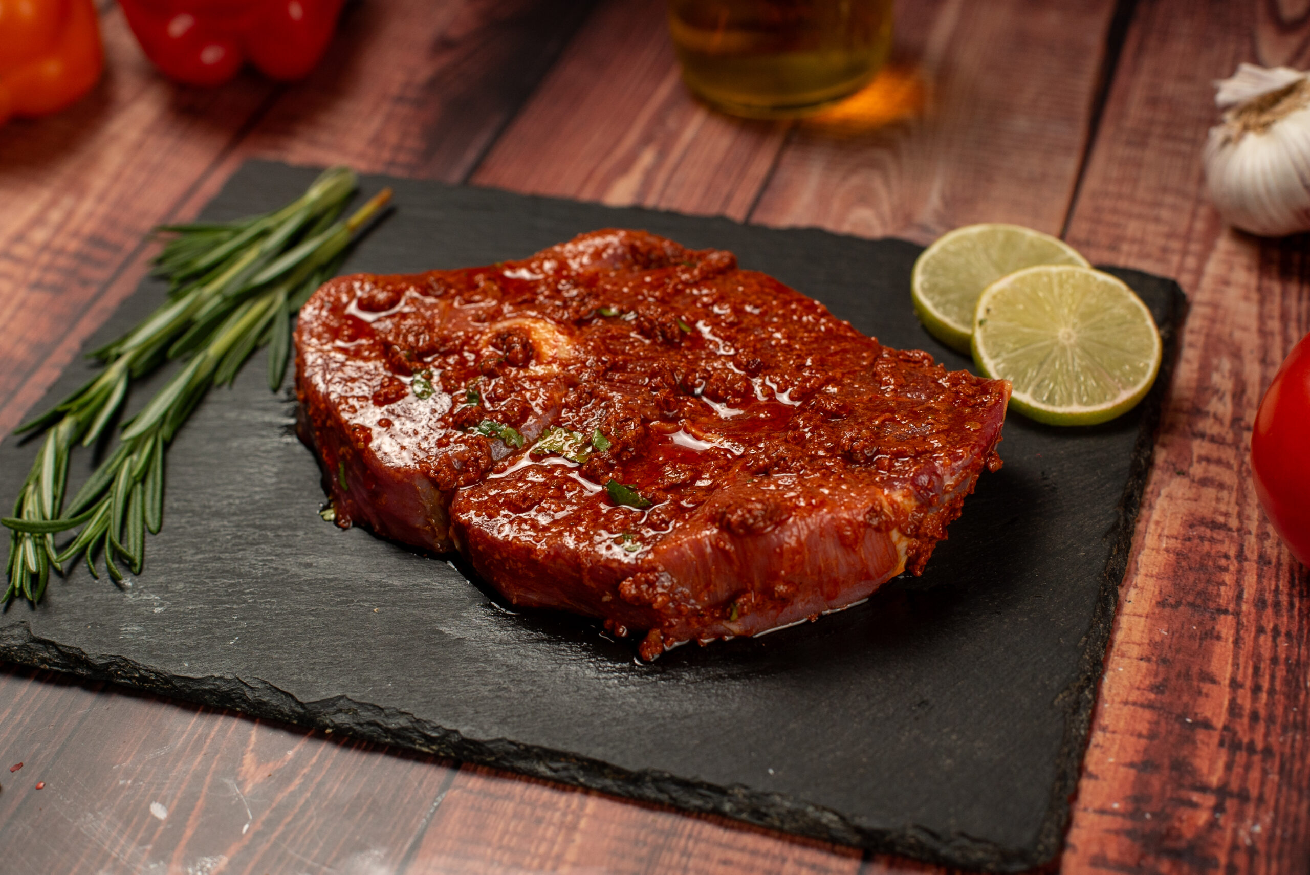 Marinated steak on slate with rosemary, lime slices, and tomatoes on a wooden table, ready for grilling.