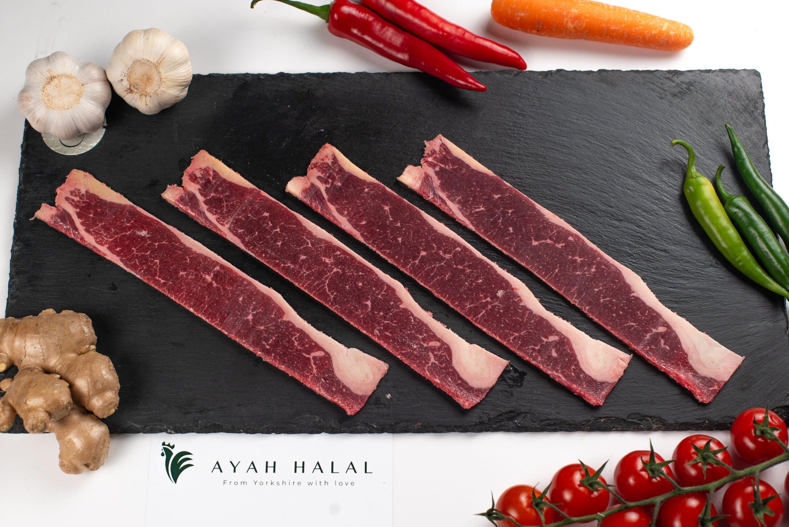 Raw halal beef strips with marbling on a slate board, fresh butcher-cut meat with garlic, chillies, ginger, and tomatoes, Ayah Halal branding visible