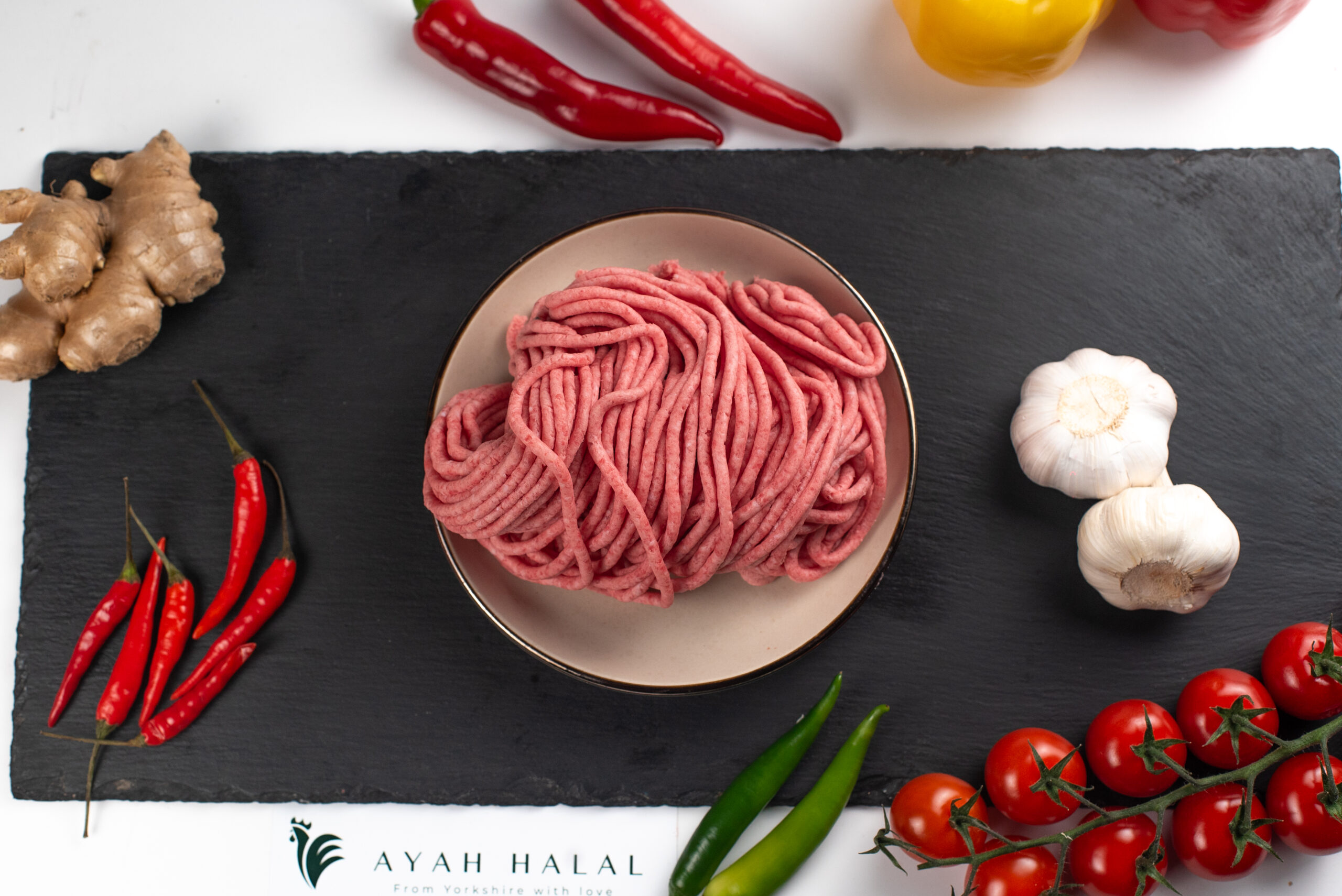 Raw ground beef mince in a bowl on a black slate board, fresh finely minced red meat with visible strands, surrounded by garlic bulbs, red and green chili peppers, cherry tomatoes, ginger root, and vegetables, with “Ayah Halal” branding visible for food preparation and cooking concept