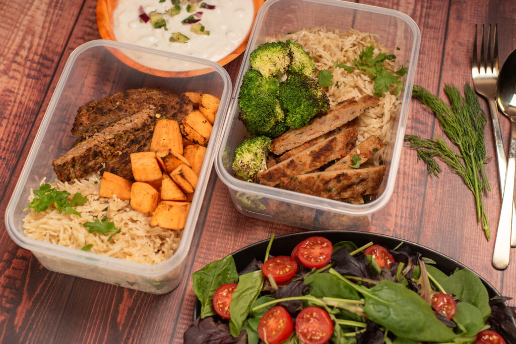 Healthy meal prep containers with basmati rice, grilled meat patties, roasted sweet potatoes, broccoli, fresh salad, and cucumber yogurt raita on rustic wooden table.