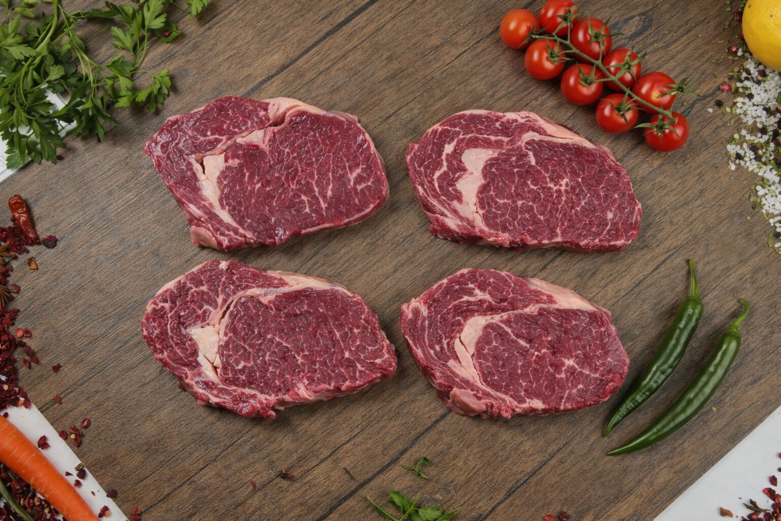 Raw beef ribeye steaks with rich marbling arranged on a wooden surface, fresh red meat cuts surrounded by cherry tomatoes, green chili peppers, herbs, spices, and vegetables for premium steak preparation and grilling concept